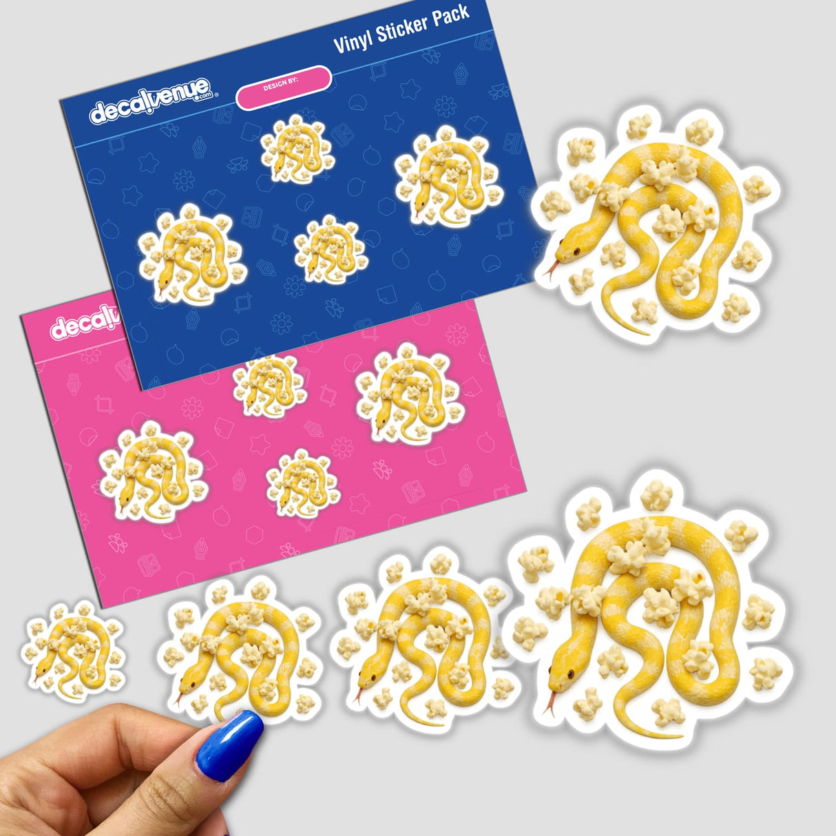 Vinyl sticker sheet titled Yellow and White Snake with Popcorn, featuring die-cut stickers of a coiled snake amidst popcorn. Displayed on blue and pink backing cards, with a hand showing the smallest size.