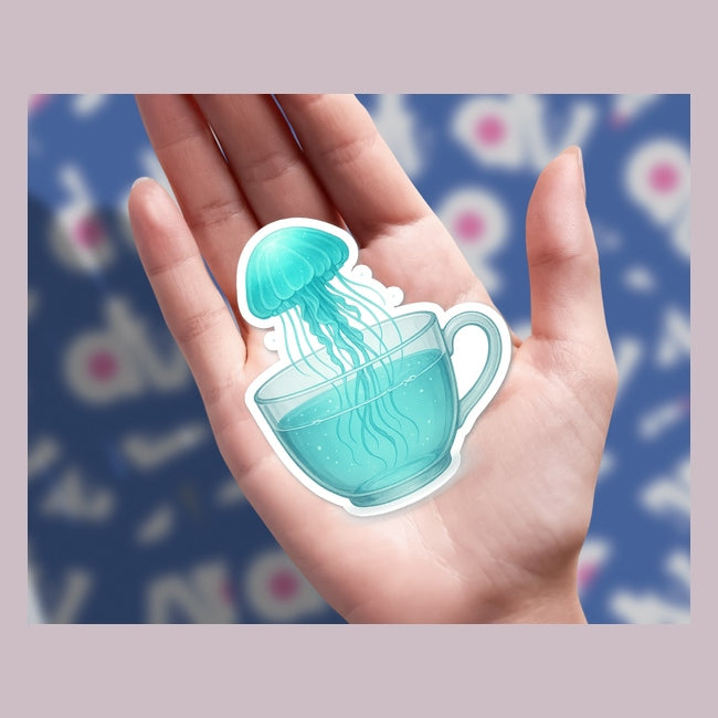 Open palm holds a die-cut illustration titled Whimsical Jellyfish Floating in a Teacup, depicting a translucent teal jellyfish seemingly drifting into a water-filled teacup, capturing Decal Venue's unique sticker artistry.